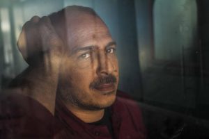 A man looks through a window with one hand resting on his head.